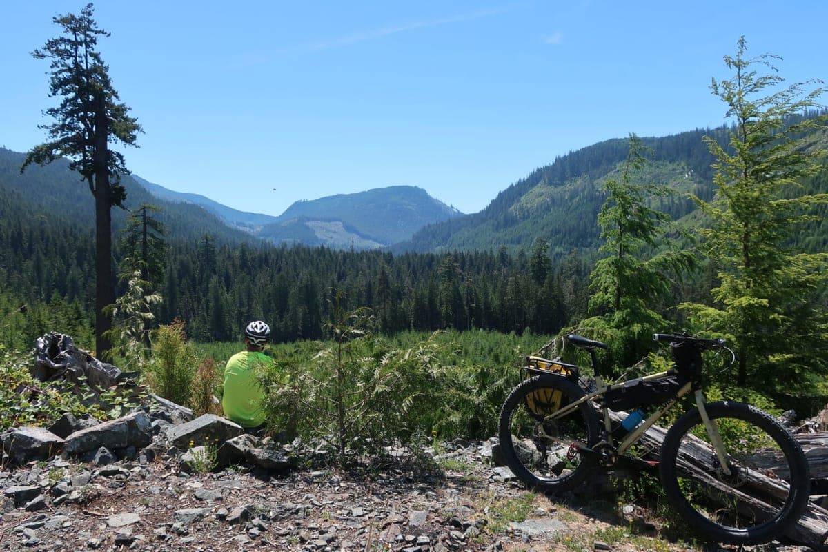 A gravel rider resting above a forested valley with a loaded bikepacking rig beside the trail